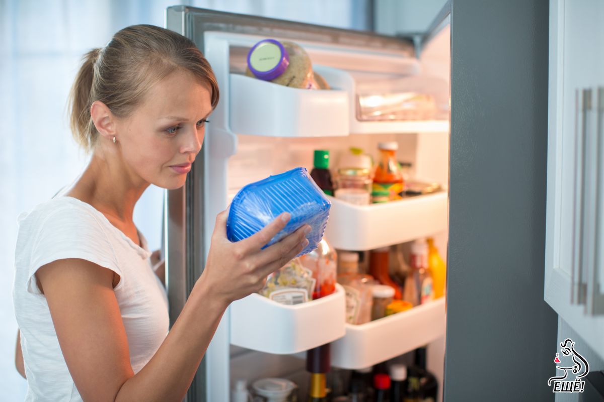 девушка стоит перед открытым холодильником и держит в руках контейнер с едой