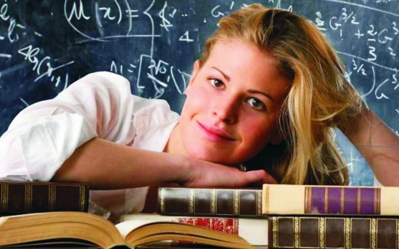 улыбающаяся девушка опирается на стопку книг на фоне исписанной формулами школьной доски