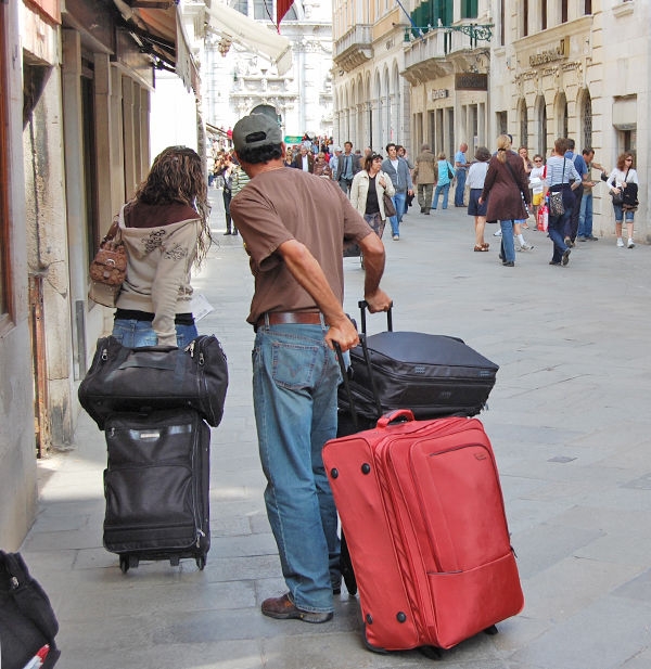 мужчина и женщина тащат и катят по узкой улице несколько огромных сумок и чемоданов