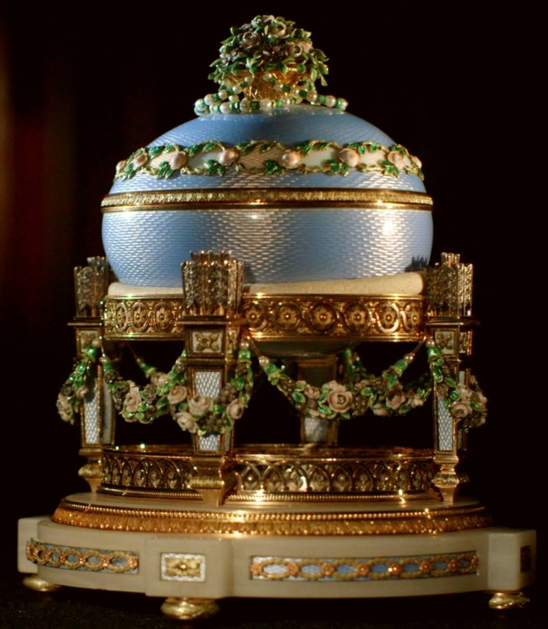 Яйцо Фаберже “Любовные трофеи” голубого цвета на золотой подставке, на темном фоне