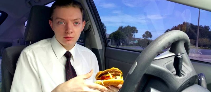 Парень при галстуке в салоне машины с гамбургером в руках