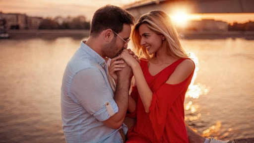 парень целует руки девушки возле реки на закате